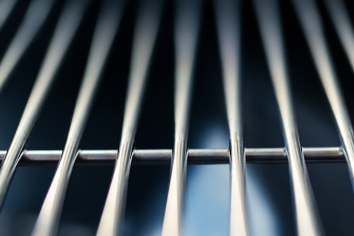 Metallic rods aligned parallel to each other, likely part of a grill or rack. The background is blurred, emphasizing the shiny texture and reflective quality of the rods.