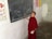 A young person in a red robe is standing by a chalkboard with writings and drawings, such as 'Live to love' and a heart shape. The wall in the background features colorful educational posters with images of flowers and anatomical diagrams.