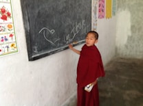 A young person in a red robe is standing by a chalkboard with writings and drawings, such as 'Live to love' and a heart shape. The wall in the background features colorful educational posters with images of flowers and anatomical diagrams.