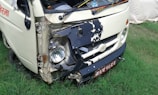A small white vehicle has damage to its front left side, with the bumper and headlight area visibly crushed and exposed. The vehicle is parked on grass, and there is some rust around the damaged area. A license plate with non-English characters is visible.