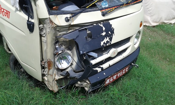 A small white vehicle has damage to its front left side, with the bumper and headlight area visibly crushed and exposed. The vehicle is parked on grass, and there is some rust around the damaged area. A license plate with non-English characters is visible.