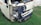 A small white vehicle has damage to its front left side, with the bumper and headlight area visibly crushed and exposed. The vehicle is parked on grass, and there is some rust around the damaged area. A license plate with non-English characters is visible.