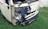 A small white vehicle has damage to its front left side, with the bumper and headlight area visibly crushed and exposed. The vehicle is parked on grass, and there is some rust around the damaged area. A license plate with non-English characters is visible.