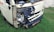 A small white vehicle has damage to its front left side, with the bumper and headlight area visibly crushed and exposed. The vehicle is parked on grass, and there is some rust around the damaged area. A license plate with non-English characters is visible.