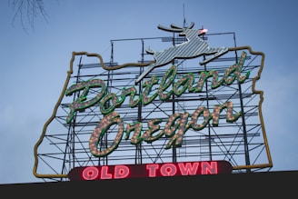 A large neon sign displays the words 'Portland Oregon' in green lettering with white highlights, set against a backdrop of a metal framework. Below, the words 'Old Town' are illuminated in red. At the top of the sign, a white outlined deer is visible.