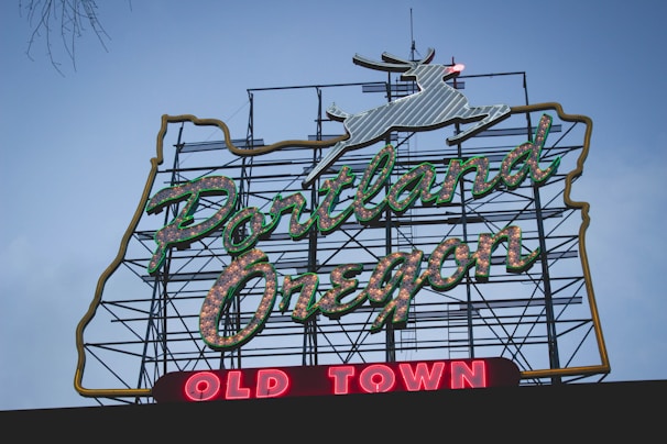 A large neon sign displays the words 'Portland Oregon' in green lettering with white highlights, set against a backdrop of a metal framework. Below, the words 'Old Town' are illuminated in red. At the top of the sign, a white outlined deer is visible.