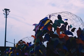 An artistic installation featuring colorful bird-shaped objects attached to wire structures. A street lamp and the outline of another wire structure are visible against a cloudy, pastel sky in the background.