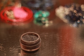 A small container of dark, glittery makeup or cosmetic powder is in focus on a reflective surface. The background is artistically blurred with colorful bokeh effect, including hues of red, green, and blue.