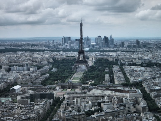 An aerial view of Paris featuring a large tower in the center, surrounded by numerous historical buildings and modern skyscrapers in the background. The scene is overcast, with cloudy skies casting a subdued light over the city, highlighting the blend of greenery and urban architecture.