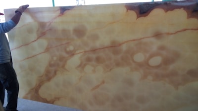 A person stands next to a large slab of translucent material with a mix of beige and brown hues, featuring organic, cloud-like patterns. The material is likely onyx, showcasing natural marbling and fluid patterns.