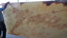 A person stands next to a large slab of translucent material with a mix of beige and brown hues, featuring organic, cloud-like patterns. The material is likely onyx, showcasing natural marbling and fluid patterns.