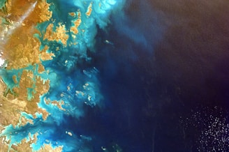 A satellite image showing a coastal area with intricate patterns of land and water. The land appears arid and brown, while the water is a rich mix of blues and turquoise.