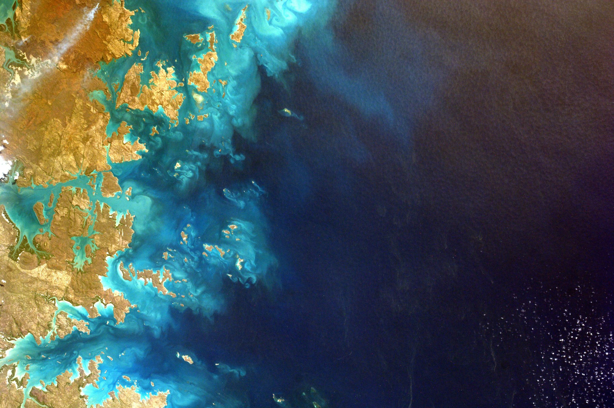 A picture of earth from space, showing coastline and light blue waters on the left, darker water on right