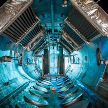 An industrial or scientific chamber with a cylindrical interior. The walls and ceiling are lined with metallic structures, vents, and panels, suggesting a complex technological or engineering environment. Central to the space are large, vertically oriented metallic structures, with cables and pipes running along the surfaces. The lighting casts a teal hue, enhancing the high-tech and futuristic appearance.