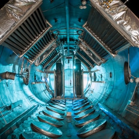 An industrial or scientific chamber with a cylindrical interior. The walls and ceiling are lined with metallic structures, vents, and panels, suggesting a complex technological or engineering environment. Central to the space are large, vertically oriented metallic structures, with cables and pipes running along the surfaces. The lighting casts a teal hue, enhancing the high-tech and futuristic appearance.