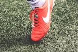 A close-up view of a red soccer cleat with white laces and a white logo on green artificial turf. The shoe is worn by a person whose sock is visible above the shoe.
