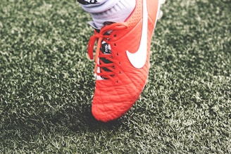 A close-up view of a red soccer cleat with white laces and a white logo on green artificial turf. The shoe is worn by a person whose sock is visible above the shoe.