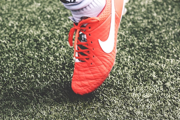 A close-up view of a red soccer cleat with white laces and a white logo on green artificial turf. The shoe is worn by a person whose sock is visible above the shoe.