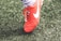 A close-up view of a red soccer cleat with white laces and a white logo on green artificial turf. The shoe is worn by a person whose sock is visible above the shoe.