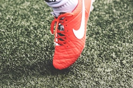 A close-up view of a red soccer cleat with white laces and a white logo on green artificial turf. The shoe is worn by a person whose sock is visible above the shoe.