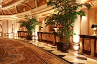 A lavishly decorated interior with ornate ceilings and marble floors. Tall potted plants line the corridor, and elegant lamps sit on intricately designed wooden tables. The lighting is warm, adding a luxurious feel to the space.