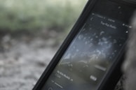 A smartphone displaying a music streaming app with a track by J. Cole playing. The screen shows album art with a blurred, muted background that appears to be outdoors.