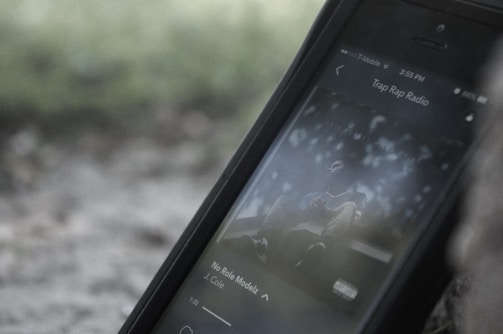 A smartphone displaying a music streaming app with a track by J. Cole playing. The screen shows album art with a blurred, muted background that appears to be outdoors.