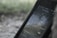 A smartphone displaying a music streaming app with a track by J. Cole playing. The screen shows album art with a blurred, muted background that appears to be outdoors.