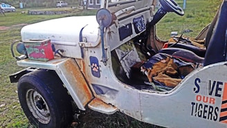A vintage, rusted white jeep is parked on a grassy area. The interior is worn and the seats are tattered. A colorful metal box is placed on the fender. The side of the jeep has a 'Save Our Tigers' sticker. The vehicle appears neglected and abandoned.