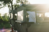 An off-road vehicle with green canvas covers is park amidst a natural setting, with trees in the background. The scene is bathed in soft, warm light, likely from the setting or rising sun. There are people in the vehicle, with one person seen adjusting something on the vehicle's side while another person is atop the vehicle.