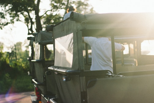 An off-road vehicle with green canvas covers is park amidst a natural setting, with trees in the background. The scene is bathed in soft, warm light, likely from the setting or rising sun. There are people in the vehicle, with one person seen adjusting something on the vehicle's side while another person is atop the vehicle.