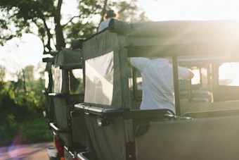 An off-road vehicle with green canvas covers is park amidst a natural setting, with trees in the background. The scene is bathed in soft, warm light, likely from the setting or rising sun. There are people in the vehicle, with one person seen adjusting something on the vehicle's side while another person is atop the vehicle.