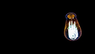 A single illuminated light bulb against a solid black background. The bulb emits a bright, white light with hints of blue and yellow. Its vintage design features a visible filament inside.
