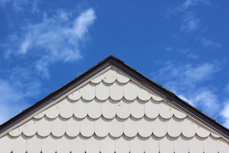 A triangular section of a white house wall with scalloped shingles is visible below a clear blue sky with scattered clouds. The shingles create a textured pattern, adding visual interest to the image.
