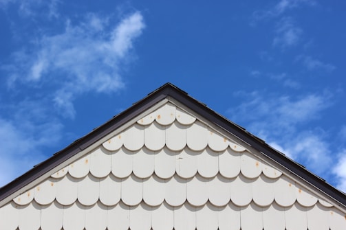 A triangular section of a white house wall with scalloped shingles is visible below a clear blue sky with scattered clouds. The shingles create a textured pattern, adding visual interest to the image.