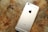A gold-colored smartphone lies face down on a wooden surface showing its back panel with a prominent logo and camera lens at the top.
