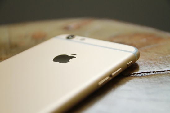 A gold-colored smartphone with a visible Apple logo lying on a wooden surface. The focus is on the top rear part of the device, highlighting its smooth texture and craftsmanship.