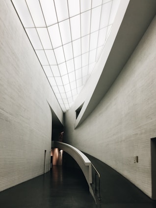 A sleek, modern architectural interior featuring clean, curved lines and a minimalistic design. The ceilings are high with a grid-like pattern of recessed lights. The walls are smooth and mostly gray, and the front view shows a ramp leading down a hallway, indicating accessibility and modernity.