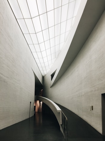A sleek, modern architectural interior featuring clean, curved lines and a minimalistic design. The ceilings are high with a grid-like pattern of recessed lights. The walls are smooth and mostly gray, and the front view shows a ramp leading down a hallway, indicating accessibility and modernity.
