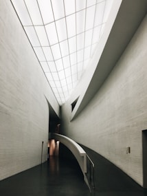 A sleek, modern architectural interior featuring clean, curved lines and a minimalistic design. The ceilings are high with a grid-like pattern of recessed lights. The walls are smooth and mostly gray, and the front view shows a ramp leading down a hallway, indicating accessibility and modernity.