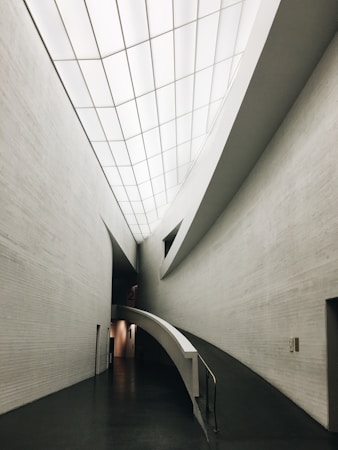 A sleek, modern architectural interior featuring clean, curved lines and a minimalistic design. The ceilings are high with a grid-like pattern of recessed lights. The walls are smooth and mostly gray, and the front view shows a ramp leading down a hallway, indicating accessibility and modernity.