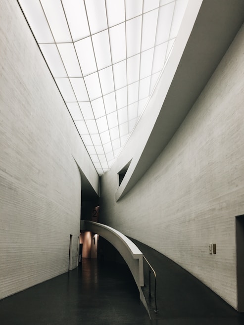 A sleek, modern architectural interior featuring clean, curved lines and a minimalistic design. The ceilings are high with a grid-like pattern of recessed lights. The walls are smooth and mostly gray, and the front view shows a ramp leading down a hallway, indicating accessibility and modernity.