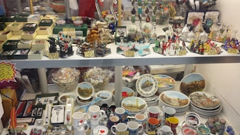A display case filled with various souvenirs including miniature sculptures, decorative plates featuring landmarks, colorful glass figurines, and a range of mugs with city themes. There are also jewelry boxes, keychains, and small replicas, arranged neatly with price tags.