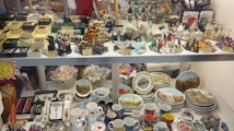 A display case filled with various souvenirs including miniature sculptures, decorative plates featuring landmarks, colorful glass figurines, and a range of mugs with city themes. There are also jewelry boxes, keychains, and small replicas, arranged neatly with price tags.