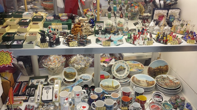 A display case filled with various souvenirs including miniature sculptures, decorative plates featuring landmarks, colorful glass figurines, and a range of mugs with city themes. There are also jewelry boxes, keychains, and small replicas, arranged neatly with price tags.
