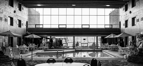 A black and white image of an indoor pool area featuring a small swimming pool surrounded by tables, chairs, and umbrellas. The area appears to have a high ceiling with wooden beams and large windows allowing natural light to enter. Several lounge chairs and potted plants are placed around the pool, creating a relaxing atmosphere. In the distance, there is a vending machine and a few people near it.