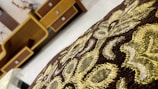 An intricate fabric with golden and brown embroidery in the foreground, with a background of wooden furniture featuring geometric designs.
