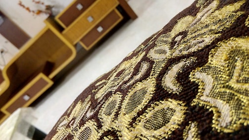 An intricate fabric with golden and brown embroidery in the foreground, with a background of wooden furniture featuring geometric designs.