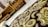 An intricate fabric with golden and brown embroidery in the foreground, with a background of wooden furniture featuring geometric designs.