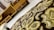 An intricate fabric with golden and brown embroidery in the foreground, with a background of wooden furniture featuring geometric designs.
